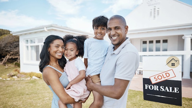 Smiling family in front of recently purchased home with 'sold' sign.