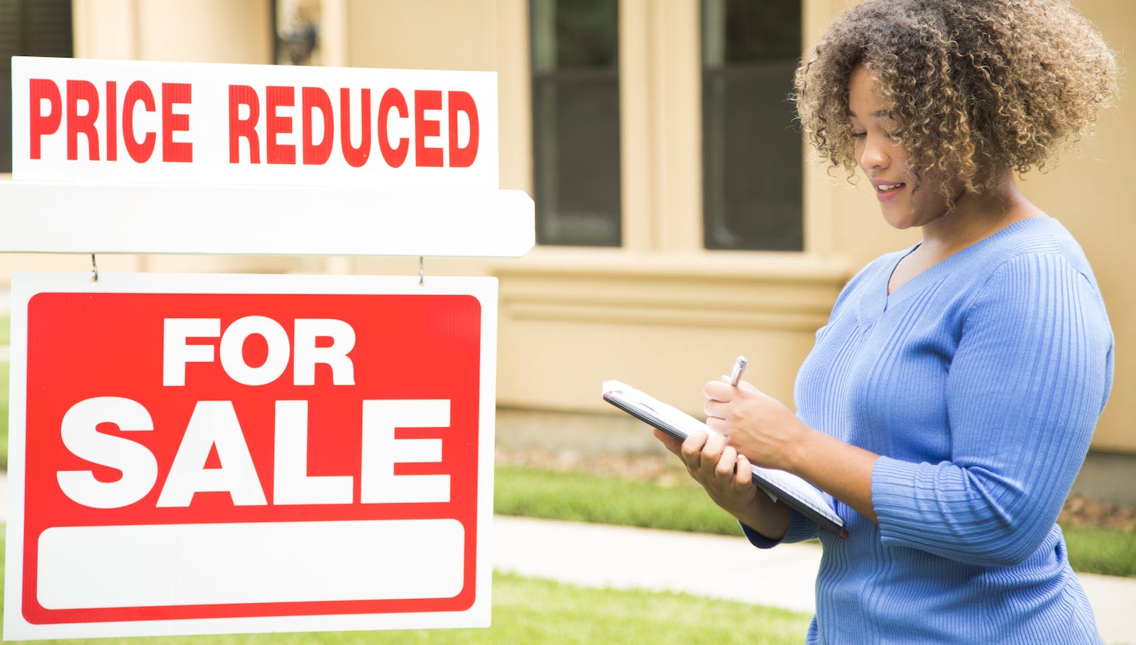 Real estate agent stands near home 'for sale' sign with 'price reduced' notice added on top