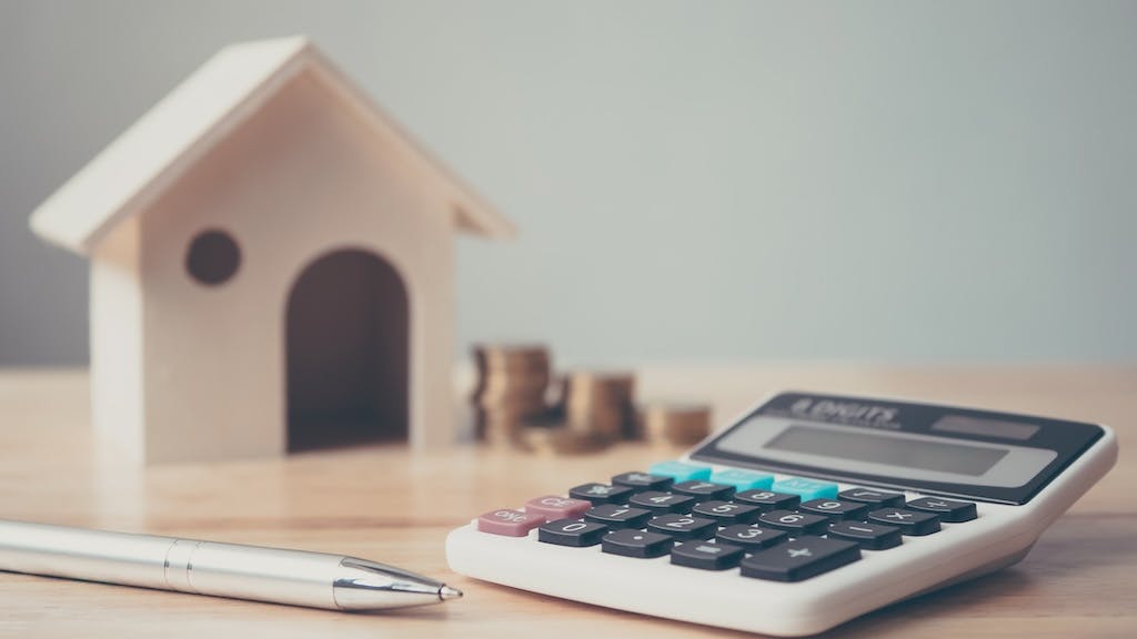 A wooden model house, calculator, pen, and stacks of coins represent the rising cost of housing, with insurance premiums making up a growing part of that cost