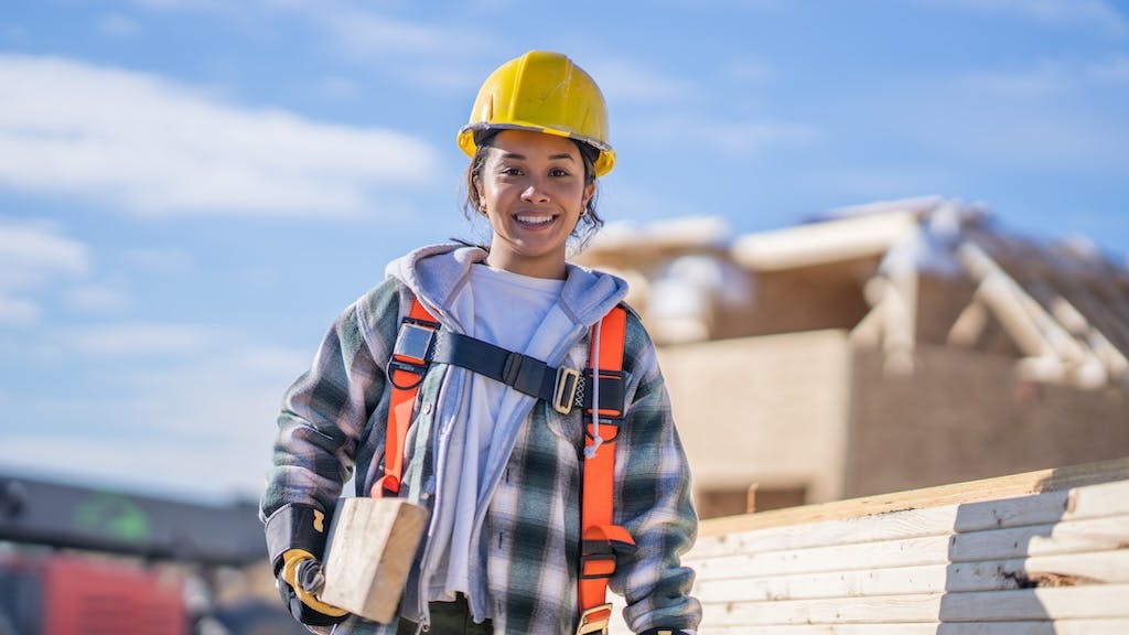 A female construction worker carries lumber through a construction site.