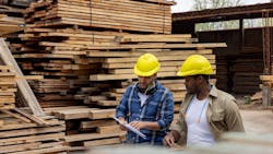 Two construction workers stand in front of stacked lumber in a lumberyard reviewing building materials prices on a clipboard Two construction workers stand in front of stacked lumber in a lumberyard reviewing building materials prices on a clipboard