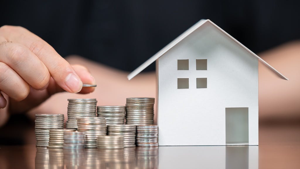 A hand stacks coins next to a model home, indicating growing home equity.