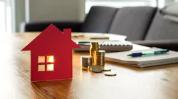A stack of coins sits behind a model of a home, indicating high home prices