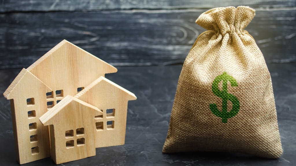 Model wooden homes sit next to large bag of money, indicating the surge in housing costs