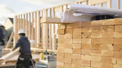 A stack of lumber with a builder working on a home in the background