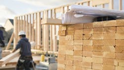 A stack of lumber with a builder working on a home in the background A stack of lumber with a builder working on a home in the background