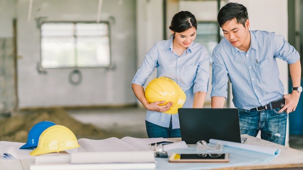 A man and a woman holding a hard hat looks over plans on a laptop at a construction site