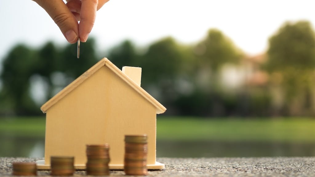 Hand places coins in stacks near model home, indicating saving for a down payment