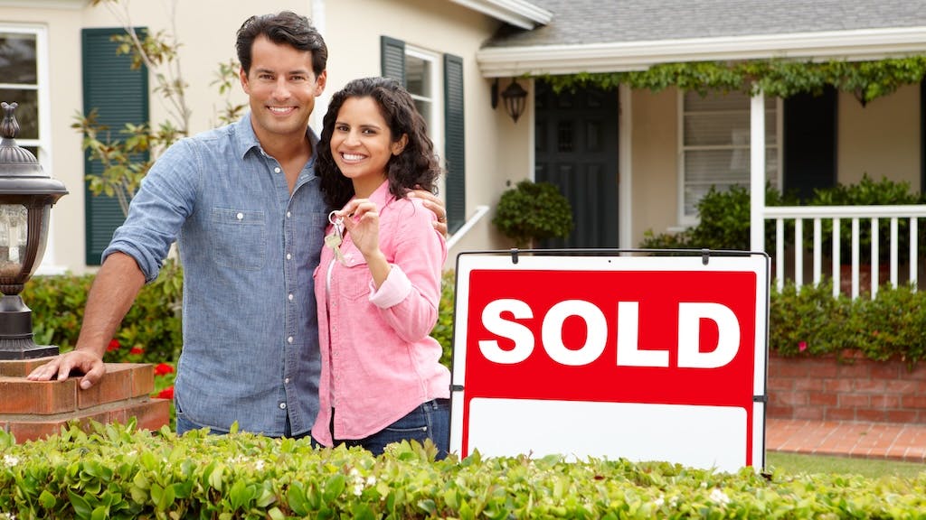 Couple holds up keys, posing next to 'sold' sign in front of their new home