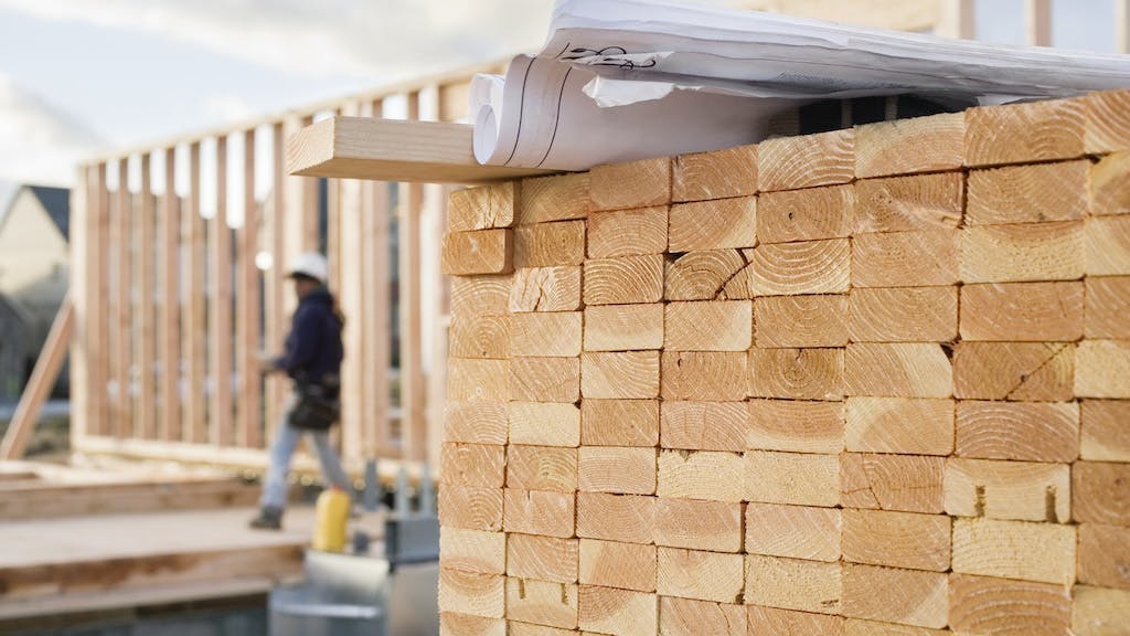 A stack of lumber in the foreground with a construction worker framing a house in the background