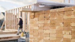 A stack of lumber in the foreground with a construction worker framing a house in the background A stack of lumber in the foreground with a construction worker framing a house in the background